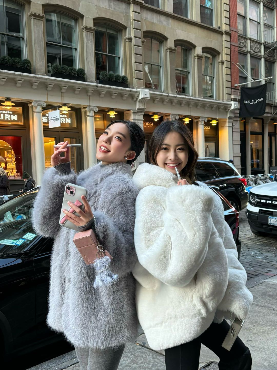 Two women in fur coats taking a selfie on a city street.